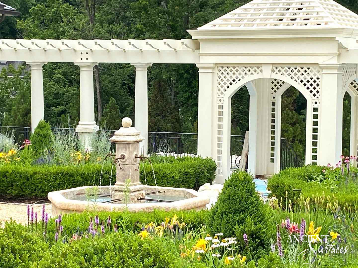 Antique limestone pillar fountain beneath a white pergola in a lush garden