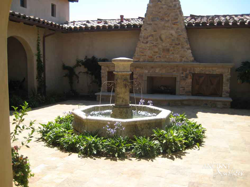 Antique limestone pillar fountain in a Mediterranean courtyard beside a stone fireplace